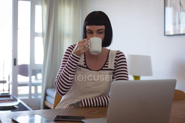 Jovem mulher tomando café enquanto usa laptop em casa — Fotografia de Stock