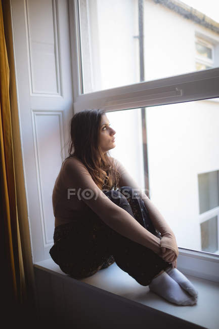 Ragionevole donna guardando attraverso la finestra mentre seduto sul davanzale della finestra a casa — Foto stock