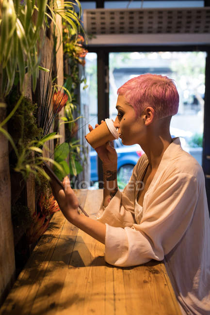 Stilvolle Frau mit rosafarbenen Haaren trinkt Kaffee, während sie im Café ein digitales Tablet benutzt. — Stockfoto