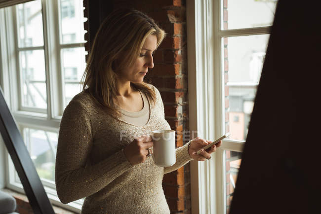 Femme utilisant un téléphone portable tout en prenant un café à la maison — Photo de stock