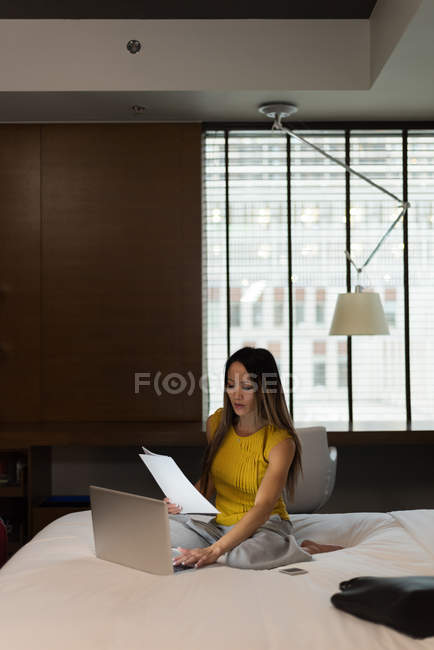 Donna d'affari seduto letto in possesso di documenti mentre si lavora sul computer portatile in hotel — Foto stock