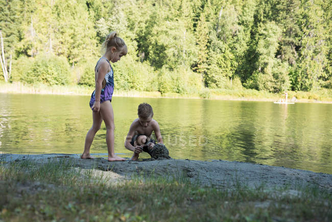 Hermano jugando con barro cerca de la orilla del río en un día soleado - foto de stock