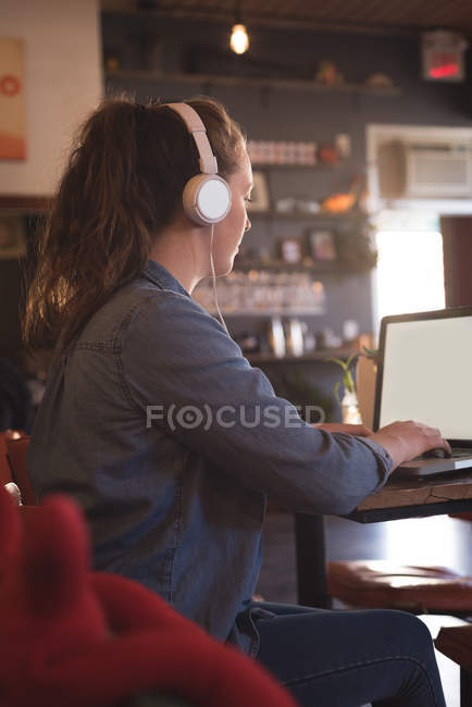 Mujer escuchando música mientras usa el portátil en la mesa en la cafetería - foto de stock