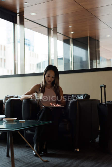 Asian businesswoman holding coffee mug while using her tablet in the lobby — Stock Photo