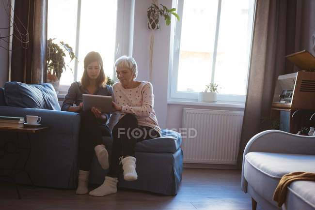 Mujer e hija mayores usando una pestaña mientras están sentadas en el sofá durante el día - foto de stock