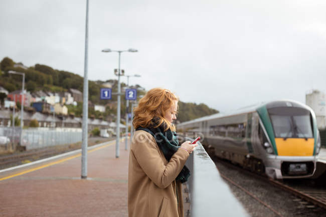 Jovem mulher de pé na plataforma ferroviária usando seu telefone celular em um dia chuvoso — Fotografia de Stock