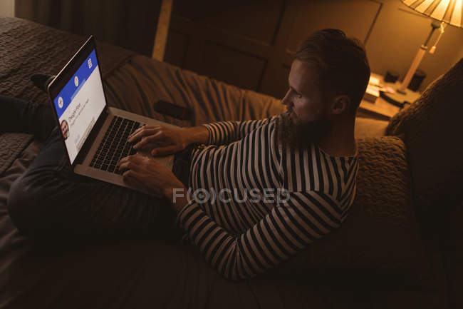Человек сидит на кровати в темной комнате, используя свой ноутбук дома — стоковое фото