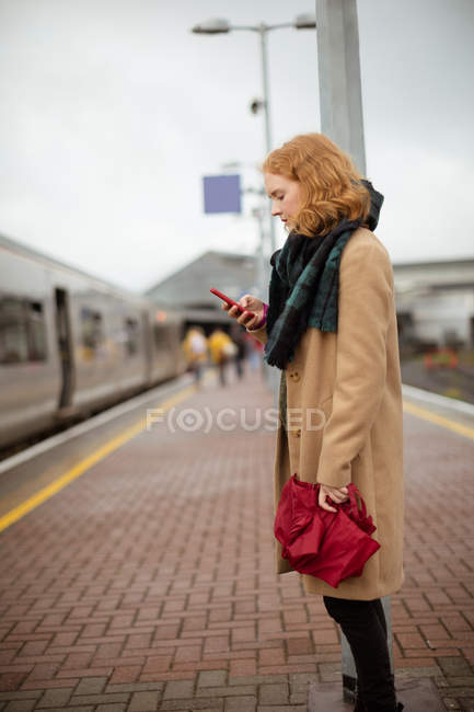 Junge Frau steht an einem regnerischen Tag mit ihrem Handy auf dem Bahnsteig — Stockfoto