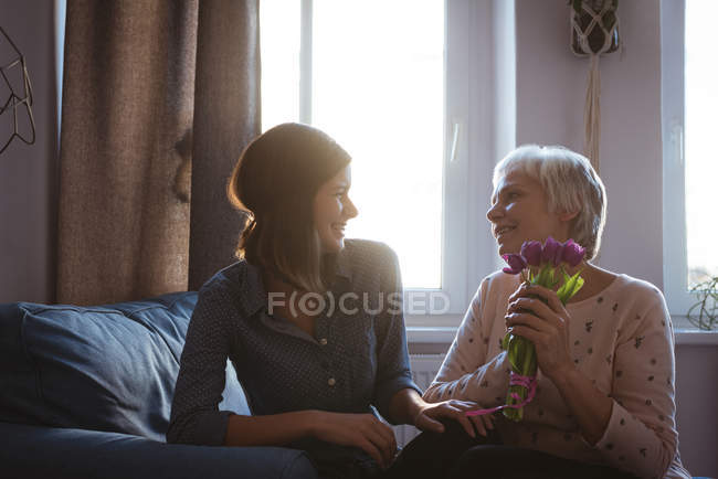 Hija dando flores de mujer mayor mientras está sentada en un sofá en la sala de estar en casa - foto de stock