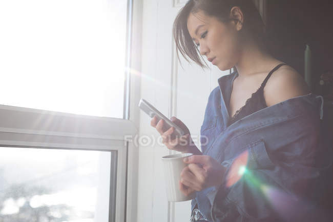 Женщина пьет кофе дома, пользуясь мобильным телефоном — стоковое фото