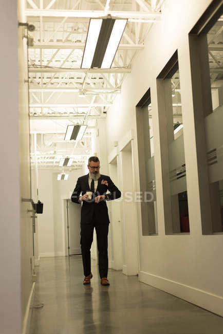 Business executive looking at smartwatch while having coffee in office corridor — Stock Photo