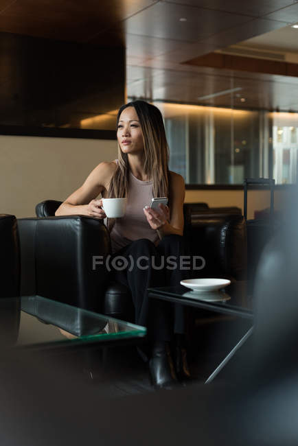 Азиатская деловая женщина смотрит в сторону, держа кофейную кружку и мобильный телефон в холле — стоковое фото