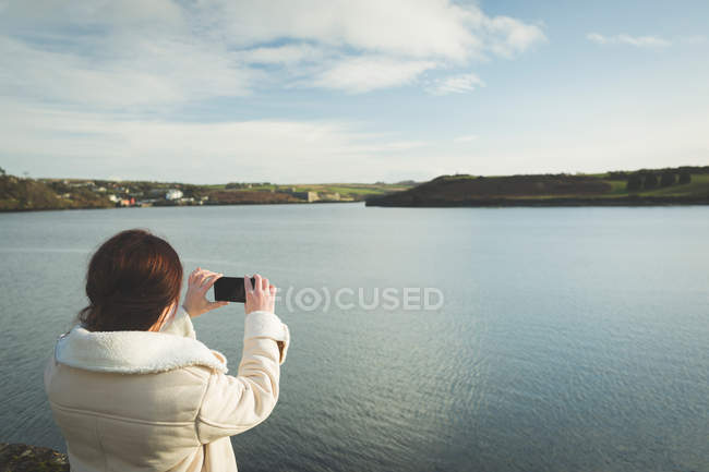 Mujer tomando fotos con teléfono móvil cerca de la orilla del río durante el atardecer . - foto de stock