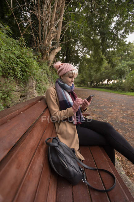 Jeune femme assise sur le banc en utilisant son téléphone portable au parc — Photo de stock