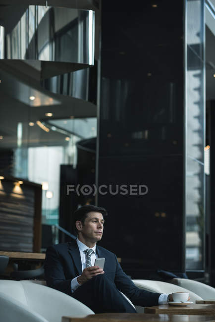 Pensativo hombre de negocios usando el teléfono móvil mientras toma café en el vestíbulo del hotel - foto de stock