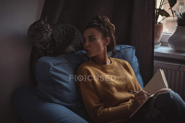 Женщина дуется на своего домашнего кота во время записи в дневнике дома — стоковое фото