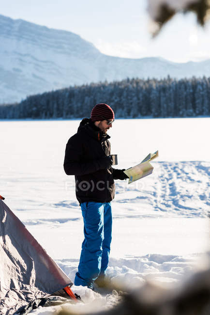 Touriste masculin regardant la carte tout en prenant un café dans un paysage enneigé par tente . — Photo de stock