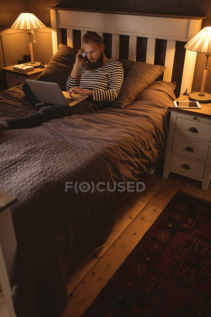 Hombre acostado en la cama hablando por teléfono mientras usa el ordenador portátil en casa - foto de stock