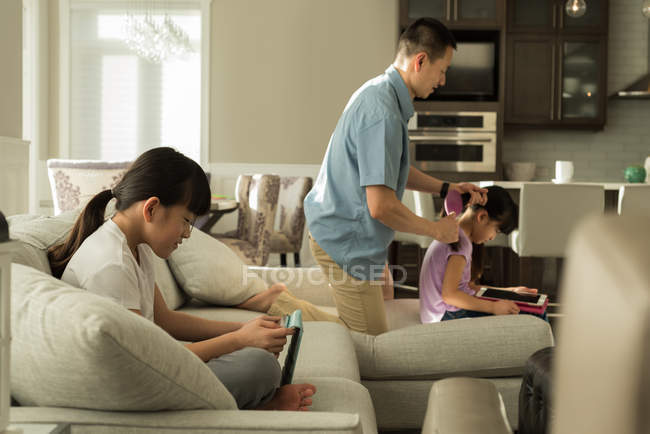 Mädchen nutzt digitales Tablet, während der Vater zu Hause die Haare seiner Töchter kämmt — Stockfoto