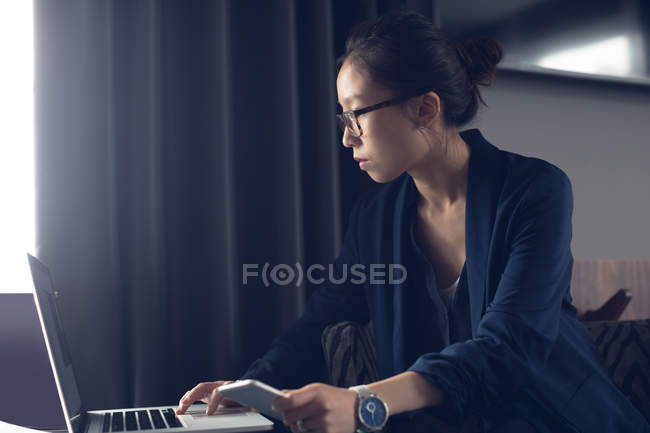 Жінка тримає мобільний телефон, використовуючи ноутбук за столом у готельному номері — стокове фото