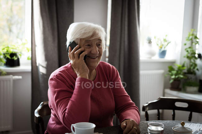 Усміхнена старша жінка розмовляє по мобільному телефону у вітальні вдома — стокове фото