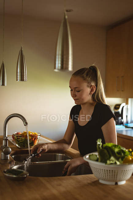 Chica de pie en la cocina y lavando pelador bajo el agua del grifo en casa . - foto de stock