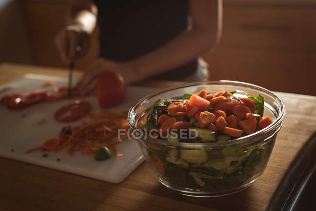 Primer plano de cuenco de verduras y chica cortando tomate en la cocina . - foto de stock