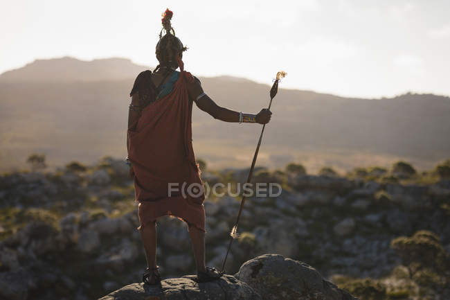 Вид ззаду чоловіка Масаї, що стоїть з палицею на скелі в сільській місцевості — стокове фото