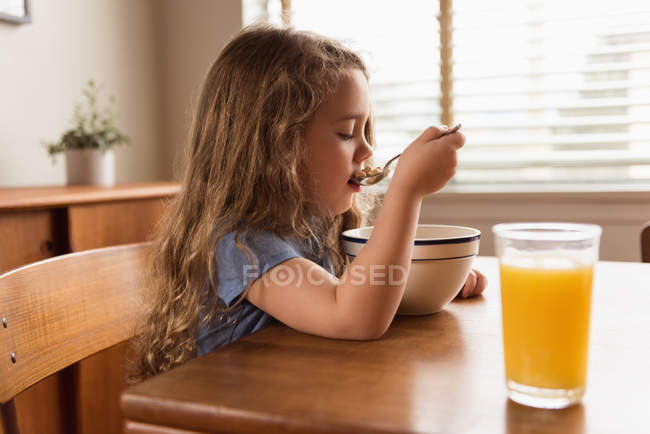 Chica desayunando cereal y jugo en la mesa en casa - foto de stock