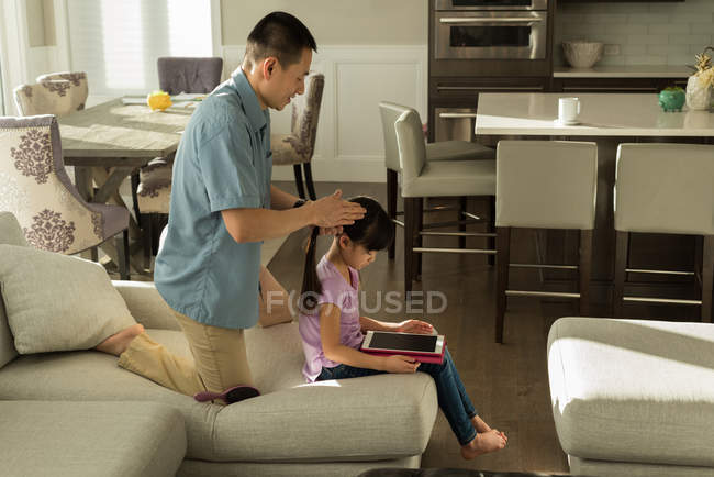 Chica usando tableta digital mientras padre peinando su cabello en casa - foto de stock