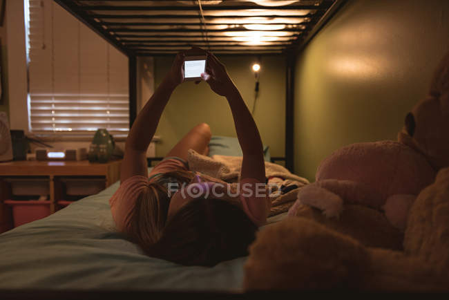 Menina deitada na cama e usando telefone celular em casa . — Fotografia de Stock