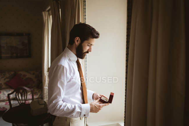 Novio sosteniendo un anillo de bodas cerca de la ventana en casa - foto de stock