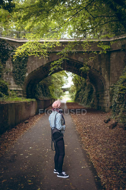 Вид сзади девушки в теплой одежде, стоящей на дорожке с рюкзаком в парке — стоковое фото