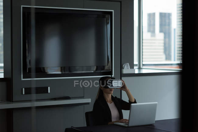 Mujer de negocios con auriculares de realidad virtual mientras trabaja en el ordenador portátil en la oficina - foto de stock