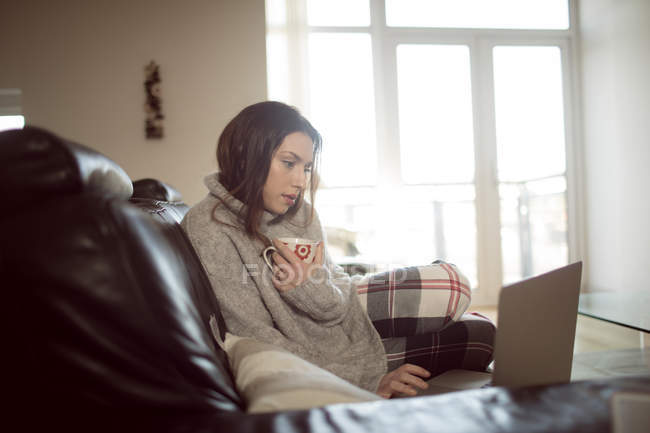 Mujer sosteniendo café mientras usa el ordenador portátil en la sala de estar en casa . - foto de stock