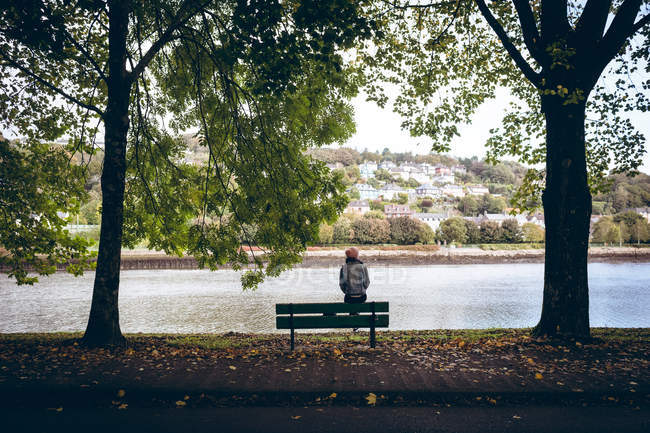 Вид спереду молода жінка сидить на лавці біля берега річки в парку — стокове фото