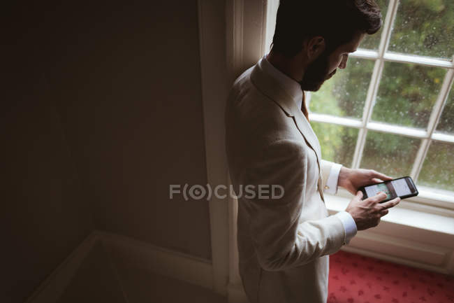 Vue grand angle du marié à l'aide de son téléphone portable près de la fenêtre — Photo de stock
