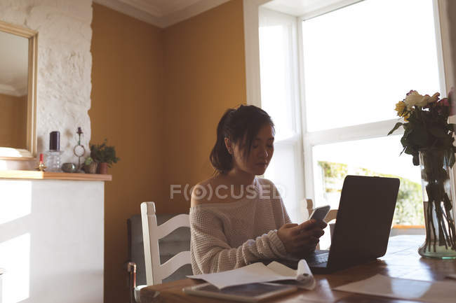 Mujer usando el teléfono móvil mientras trabaja en el ordenador portátil en casa - foto de stock