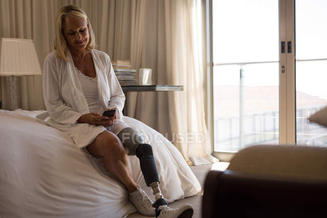 Mulher madura com perna protética usando telefone celular no quarto em casa . — Fotografia de Stock