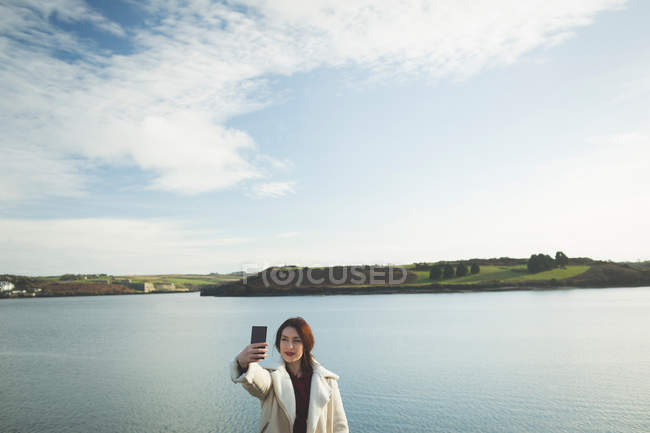 Donna che prende selfie con il telefono cellulare vicino al fiume durante il tramonto . — Foto stock