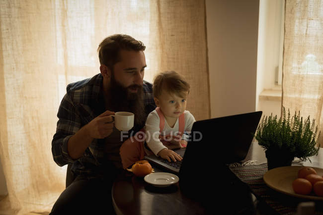 Отец пьет кофе, а сын играет дома на ноутбуке. — стоковое фото