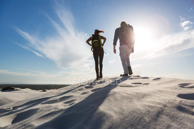 Vista trasera de pareja con mochila caminando sobre arena en un día soleado - foto de stock