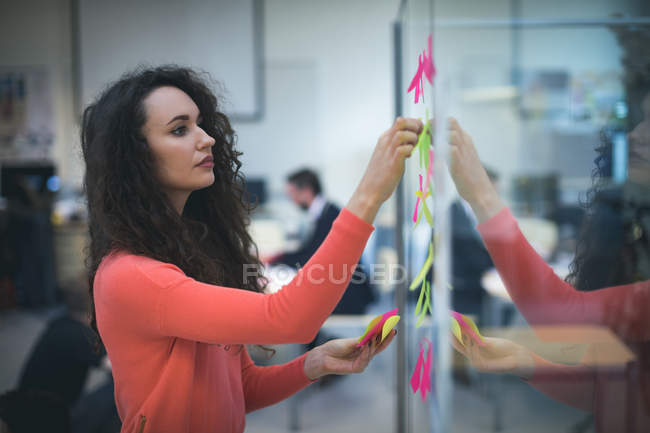 Exécutif féminin collant des notes adhésives sur le mur de verre dans le bureau — Photo de stock