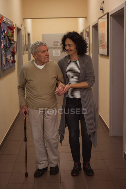 Смотритель помогает пожилому человеку во время прогулки по коридору в доме престарелых — стоковое фото