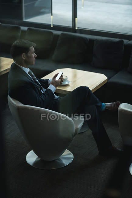 Thoughtful businessman using mobile phone while having coffee in hotel lobby — Stock Photo