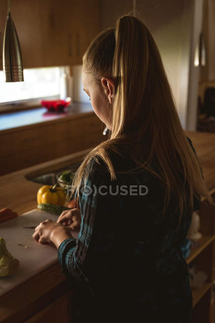 Pre-adolescent girl standing in kitchen and cutting vegetables with knife at home. — Stock Photo