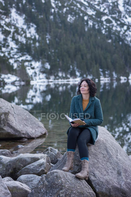 Задумчивая женщина, сидящая с книгой на берегу озера зимой — стоковое фото