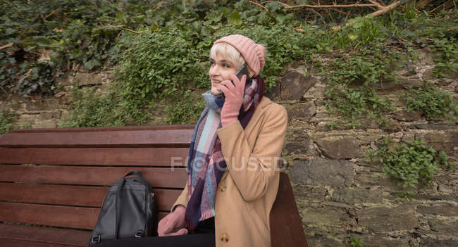 Mujer joven hablando por teléfono mientras está sentada en el banco en el parque - foto de stock