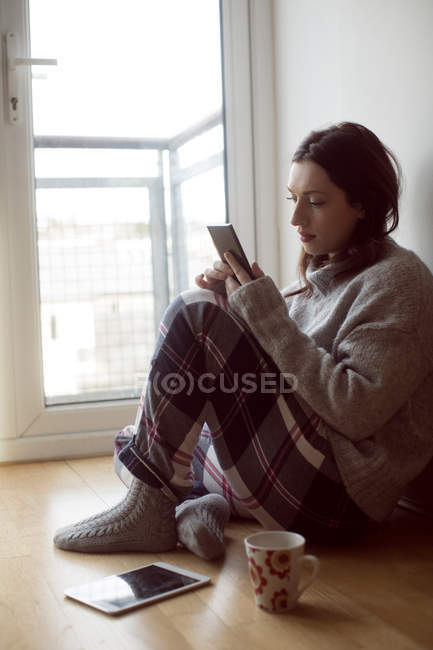 Молода жінка використовує мобільний телефон, сидячи на підлозі вдома . — стокове фото