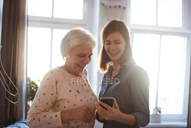 Mujer e hija mayores usando un teléfono móvil en la sala de estar en casa - foto de stock